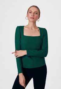 Zielony prążkowany top z długim rękawem i kwadratowym dekoltem, ozdobiony marszczonym detalem na ramionach, w połączeniu z ciemnymi spodniami. Tkanina wydaje się mieć fakturę.