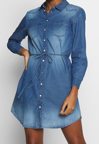 Denim-paita-mekko, jossa on nappikiinnitys edessä, kaksi rintataskua, vyötäröllä vyö ja siniharmaata sävyä, joka vaihtelee tummasta vaalennukseen. Pitkät hihat.