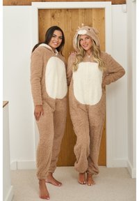 Twee vrouwen dragen bruine fleece onesies met crèmekleurige accenten. De capuchons hebben dierengezichten, met een textuur van zachte stof en ritssluitingen.