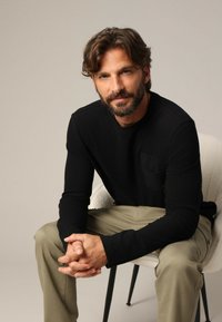 Chemise noire à manches longues avec une poche sur la poitrine, associée à un pantalon beige clair. L'individu est assis, les mains jointes sur les genoux.