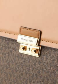 Ruskea ja beige käsilaukku, jossa on teksturoidusta kankaasta valmistettu pinta, kullanvärinen lukko ja kohokuvioitu logolevy. Yksityiskohtina on myös avainreikä.