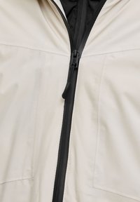 Beežist jopest lähivaade, millel on must tõmblukk ja kangast tõmblukaias, mis asub kahe esitasku vahel keskel.