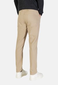 Pantalones chinos beige de corte slim, con bolsillos traseros y textura lisa, combinados con zapatillas blancas. La prenda se muestra desde la parte trasera.