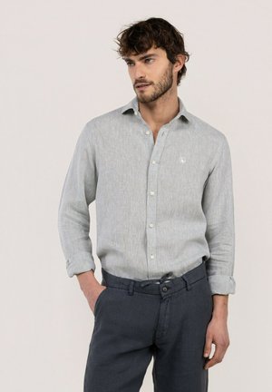 Hombre con barba que lleva una camisa gris claro de manga larga con botones y pantalones azul oscuro, de pie con una mano en el bolsillo, mirando hacia la derecha.