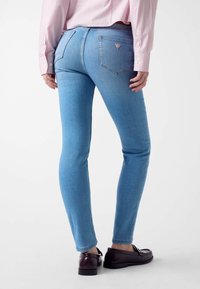 Jeans skinny bleu clair en denim, avec une taille mi-haute, des poches arrière et un petit patch logo. Texture lisse et design ajusté.