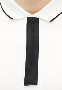 Λευκό πόλο μπλουζάκι με μαύρο περίγραμμα στο γιακά και μια κάθετη μαύρη υφασμάτινη λωρίδα με ανάγλυφη σήμανση Armani Exchange στο μπροστινό φερμουάρ.