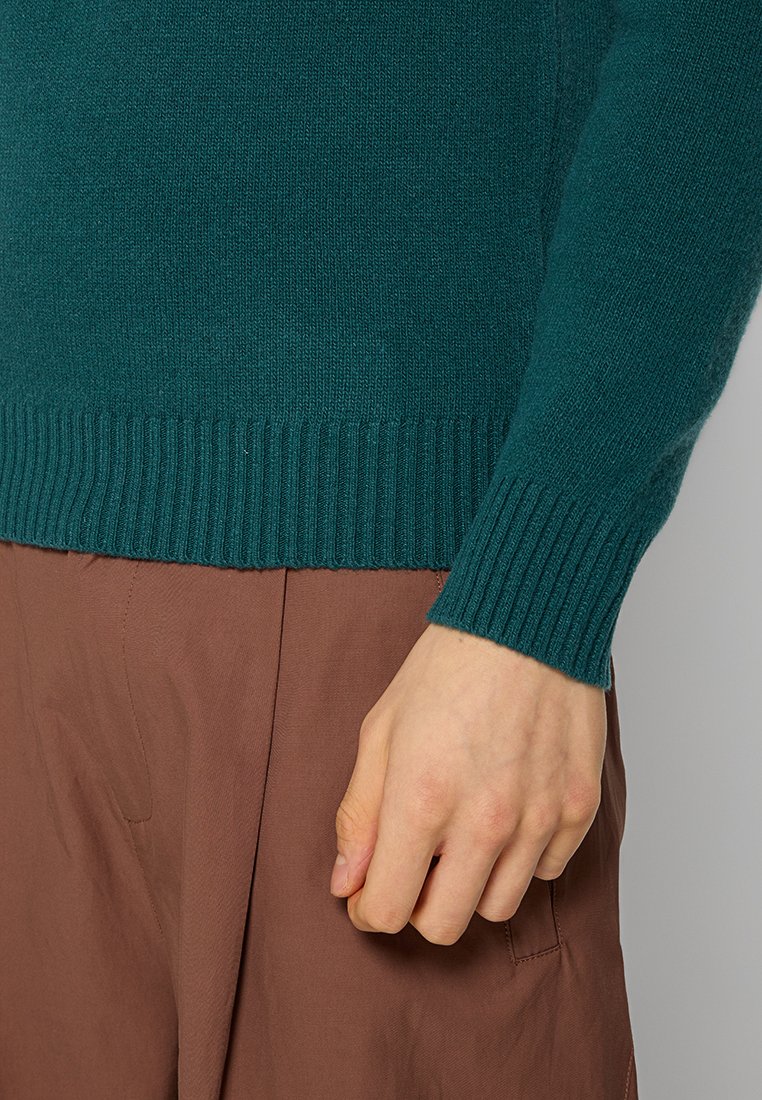 Groene gebreide trui met geribbelde manchetten, die een zachte textuur laat zien. De hand is geplaatst tegen een bruine stof, wat de kleurcontrast benadrukt.