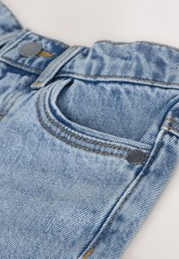 Primo piano di jeans in denim azzurro chiaro che mostrano la tasca anteriore, i dettagli delle cuciture e i rivetti metallici su sfondo bianco.
