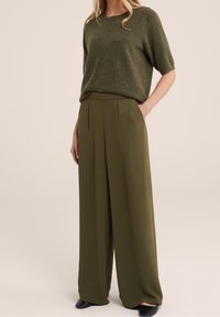 Olive green short-sleeve knit sweater paired with high-waisted, wide-leg trousers. Trousers feature pleats and textured fabric.