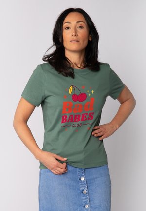 Frau mit dunklem Haar, die ein grünes T-Shirt mit roten Kirschen und dem Schriftzug "Bad Babes Club" trägt, kombiniert mit einem geknöpften Jeansrock, steht selbstbewusst.