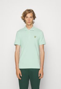 Mintgroene polo shirt van katoen, met een klassieke kraag en een knoopsluiting met drie knopen, gecombineerd met donkergroene broek. Gele logokleuraccents.