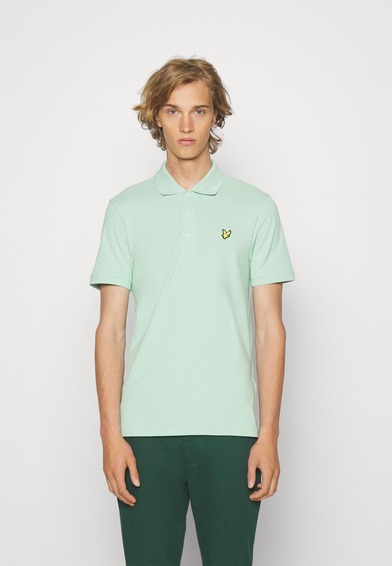 Mintgroene polo shirt van katoen, met een klassieke kraag en een knoopsluiting met drie knopen, gecombineerd met donkergroene broek. Gele logokleuraccents.