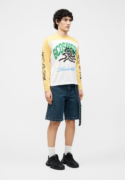 Ένας νεαρός άντρας στέκεται φορώντας ένα μακρυμάνικο πουκάμισο κίτρινο και άσπρο με πράσινα και μαύρα γραφικά, τζιν σορτς, λευκές κάλτσες και μαύρα αθλητικά παπούτσια.