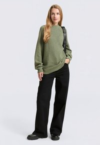 Roheline soonikkoes kõrge kaelusega kampsun, mustad laiade säärega püksid ja massiivsed mustad saapad. Modell seisab õlakotiga.