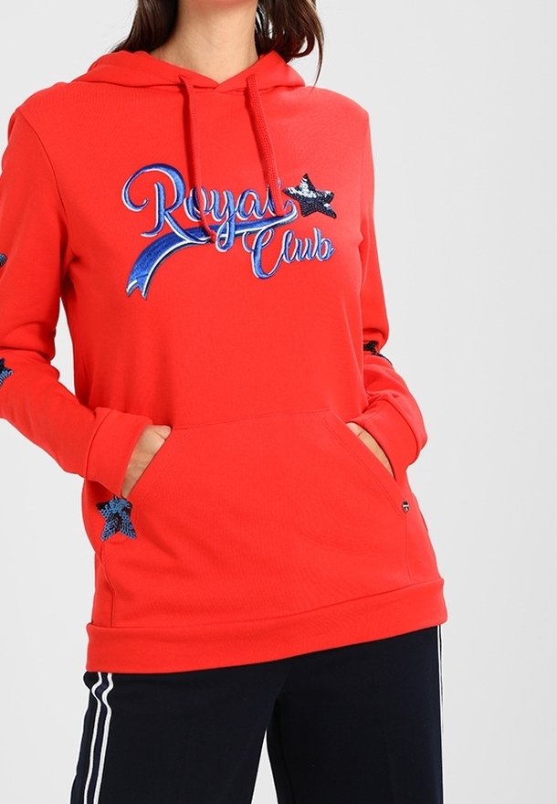 Femme portant un sweat à capuche rouge avec le texte bleu « Royal Club » et des écussons étoiles, mains dans la poche avant, associée à un pantalon noir à rayures blanches.