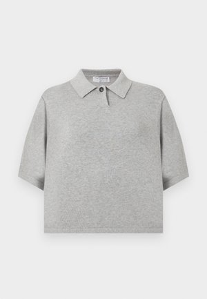 Moteris stovint su viena ranka ant klubų, vilki pilką trumpomis rankovėmis palaidinę su apykakle, baltas plačias kelnes ir juodus smailius aukštakulnius.