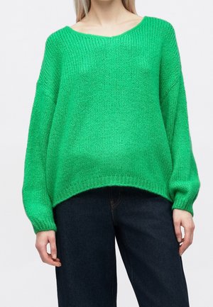 Femme portant un pull en maille ample, vert vif, avec des épaules tombantes et un jean taille haute bleu foncé, debout devant un fond blanc.