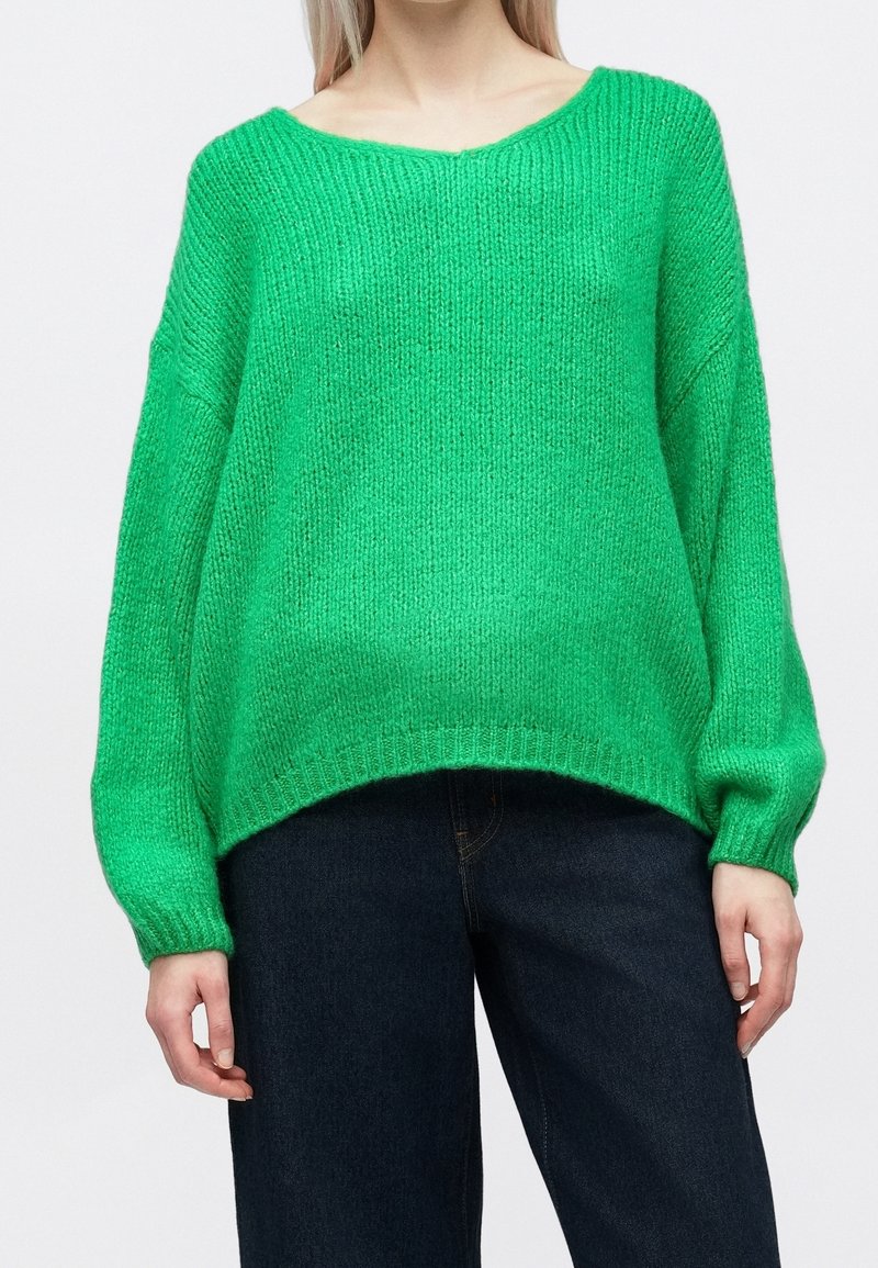 Femme portant un pull en maille ample, vert vif, avec des épaules tombantes et un jean taille haute bleu foncé, debout devant un fond blanc.