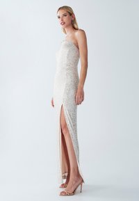 Sandy beige strapless mekko, joka ulottuu lattiaan ja on koristeltu paljeteilla, sisältää reiden korkeuden halkion ja tiukan siluetin. Yhdistettynä ihonvärisiin sandaaleihin.