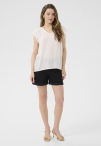 Blouse blanche légère à manches courtes avec un col en V, assortie à un short noir. Le mannequin porte des sandales à talons jaunes.