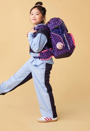Young girl wearing blue and black outfit, standing on one leg, carrying a large purple and pink patterned backpack.