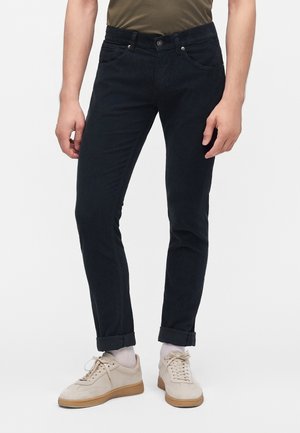Man draagt smalle zwarte broek met omslag bij de enkels, witte sokken, beige sneakers en een olijfgroen overhemd zichtbaar ter hoogte van de taille.