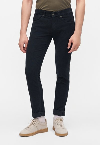 Homme portant un pantalon noir slim retroussé aux chevilles, des chaussettes blanches, des baskets beige et une chemise vert olive visible à la taille.