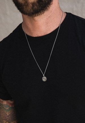 Silver pendant necklace on a thin chain, featuring a round charm with a textured surface, worn over a black shirt.