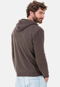 Sweat à capuche marron avec un matériau texturé et duveteux sur les manches et la capuche, présentant un ourlet côtelé et un design simple et décontracté.