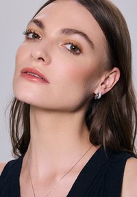 Srebrne kolczyki w kształcie obręczy z teksturowaną powierzchnią i małymi przezroczystymi kamieniami, prezentowane na modelce z długimi brązowymi włosami i naturalnym makijażem.