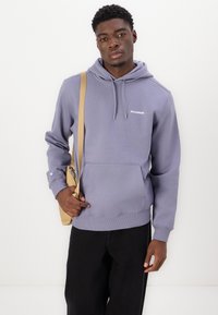 Sweatshirt à capuche violette avec une poche avant et des poignets côtelés, arborant un petit logo blanc "Salomon" sur la poitrine. Porté avec un pantalon sombre.