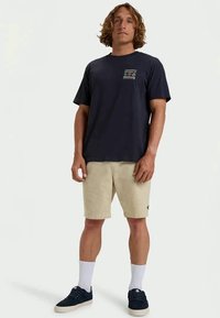 Homem em pé, vestido com uma camisola gráfica azul-marinho, calções bege, meias brancas e ténis pretos, contra um fundo branco liso.