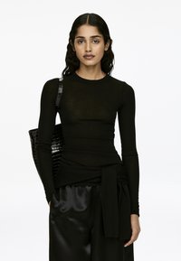 Schwarzes Langarm-Oberteil mit tailliertem Schnitt, aus einem semi-transparenten Material gefertigt. Kombiniert mit hochgeschnittenen, glänzenden schwarzen Hosen und einer strukturierten Tasche.