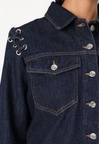 Tume denimpakiga koos hõbedaste detailide, ees olevate taskute ja dekoratiivse nöörimisega õlgadel, millel on oranžad õmblused.