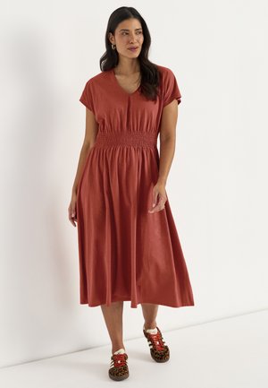 Vrouw draagt een roestkleurige midi-jurk met geplooide taille en sneakers met luipaardprint, staande tegen een witte achtergrond.