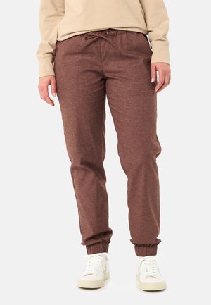 Persoon draagt bruine joggerbroek met elastische boorden, beige sweatshirt en witte sneakers, staand tegen een witte achtergrond.