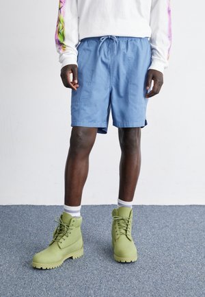 Shorts bleu clair avec une taille élastique et un cordon de serrage, associés à des bottes vertes à lacets et des chaussettes blanches montantes. Surface texturée.