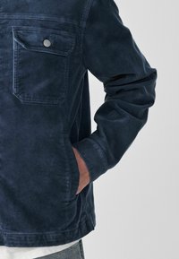 Personne portant une veste en velours côtelé bleu foncé, avec la main dans la poche latérale et un détail de poche poitrine boutonnée visible.