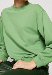 Sudadera verde con un cuello alto, puños acanalados y un corte holgado. Textura suave, con detalles de costura a lo largo de las costuras.