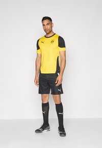 Camisa de fútbol amarilla y negra de mangas cortas, con un textura estampada, combinada con pantalones cortos negros y calcetas negras hasta la rodilla, con acentos de logotipos blancos.