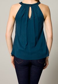 Haut sans manches bleu sarcelle avec un dos à ouverture en forme de goutte, en tissu doux, et un ourlet arrondi. Présente un col décoratif et une texture lisse.