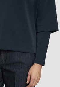Tamno plavi oversize top s okruglim izrezom, širokim rukavima i rebrastim manžetama. Glatka tekstura s vidljivim kontrastnim šavovima.