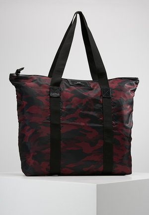 Grand sac fourre-tout avec motif camouflage rouge foncé et noir et deux longues anses en tissu noir, posé sur une surface blanche.