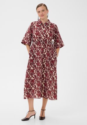 Femme portant une robe midi rouge et blanche à motifs floraux avec manches trois-quarts et des talons noirs à bride cheville, debout les mains dans les poches sur fond blanc.