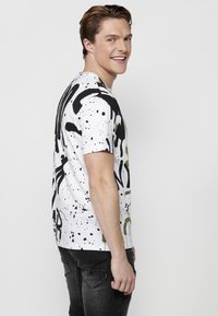 Camiseta blanca con patrones de salpicaduras abstractas en negro y verde. Cuello redondo, mangas cortas y un corte relajado. Llevada con unos vaqueros oscuros.