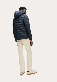 Giacca puffer blu navy con motivo a trapunte orizzontali e cappuccio regolabile, abbinata a pantaloni crema e sneakers chiare.