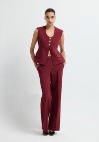 Gilet croisé bordeaux à quatre boutons dorés, associé à un pantalon à jambes larges assorti. Tissu lisse, coupe structurée, sans manches.