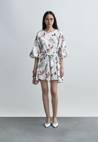 Γυναίκα που φοράει λευκό φόρεμα με floral σχέδιο και φαρδιές μανσέτες, λευκά μυτερά παπούτσια, γυαλιά και κρατά μια μικρή ροζ τσάντα, στέκεται σε γκρι δάπεδο.