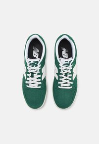 Gröna mockaskor med vita snören, som har en stor vit logotyp på sidan och en svart inredning med perforerade detaljer på ovandel.