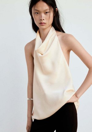 Jeune femme aux longs cheveux foncés portant un top drapé sans manches crème et un pantalon noir, posant avec une main sur la hanche sur fond uni.